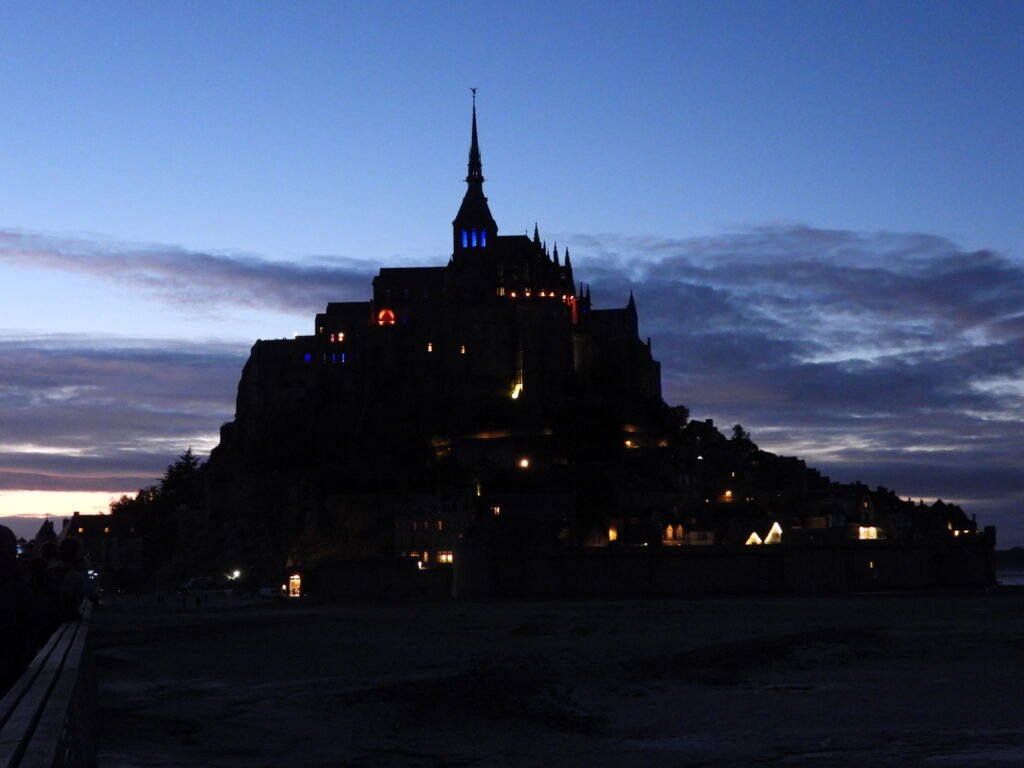Mont Saint-Michel illuminé la nuit, France. Édifices historiques et ciel nuageux en arrière-plan.