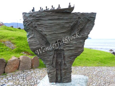 Sculpture représentant une figure humaine et des poissons, située à Elduvik, Îles Féroé, avec une vue sur la mer et des collines verdoyantes.