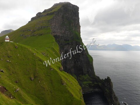 Un phare blanc se dresse sur une falaise verte à Trollanes, aux îles Féroé, surplombant l'océan et les montagnes environnantes.