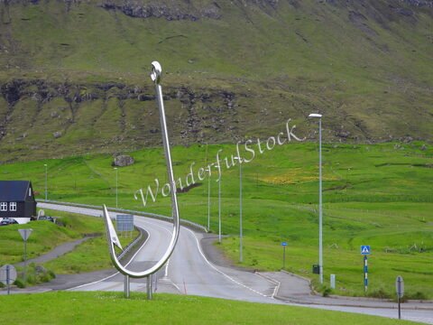Un grand crochet en métal se dresse près d'une route à Klaksvik, aux îles Féroé, avec des collines verdoyantes en arrière-plan.