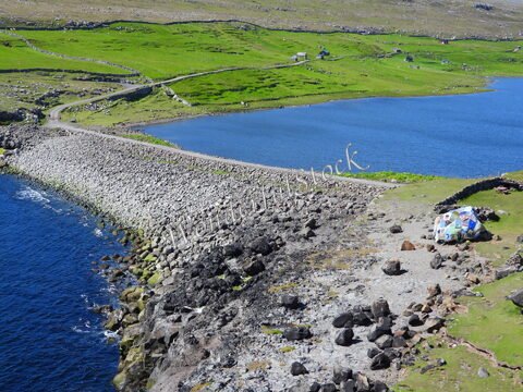 Nordradalur, Îles Féroé, présente une route sinueuse bordée de pierres et un lac entouré de collines verdoyantes.