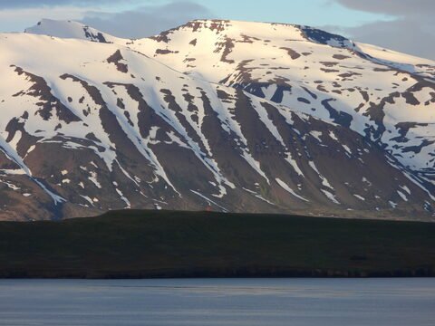 Montagnes enneigées en Islande, avec des pentes abruptes et un ciel nuageux au-dessus d'un plan d'eau calme.