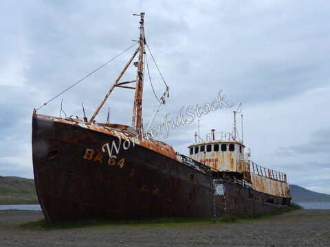 Un bateau de pêche rouillé est échoué sur le rivage en Islande. Des montagnes et un ciel nuageux sont visibles en arrière-plan.