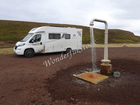 Un camping-car près d'une source d'eau en Islande. Le sol est en terre et des collines verdoyantes se trouvent en arrière-plan.