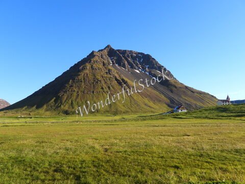 Mont verdoyant dominant Bolungarvik, Islande, avec des maisons blanches à proximité et un ciel bleu dégagé.