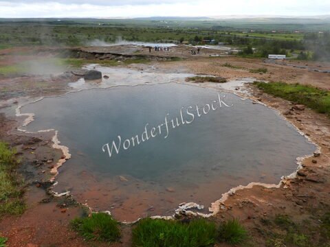 Geysir en Islande, avec un bassin d'eau et des zones de vapeur, entouré de verdure et de paysages naturels.