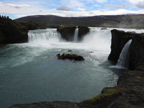 Chutes d'eau à Goðafoss, Islande, avec des rochers et des visiteurs en arrière-plan, sous un ciel nuageux.