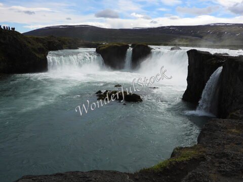 Chutes d'eau à Goðafoss, Islande, avec des rochers et des visiteurs en arrière-plan, sous un ciel nuageux.