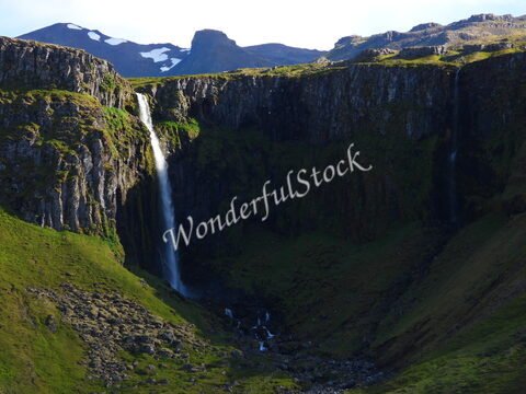 Chutes d'eau sur des falaises verdoyantes à Grundarfjörður, Islande, avec des sommets enneigés en arrière-plan.