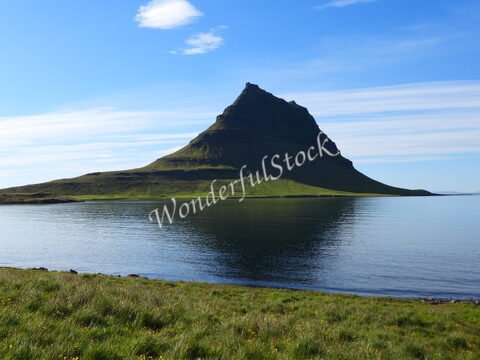 Kirkjufell en Islande, montagne conique se reflétant dans l'eau calme, entourée de verdure et sous un ciel dégagé.