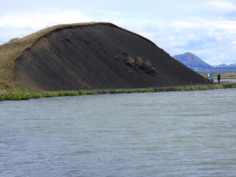 Un cône volcanique noir et une rivière se trouvent à Skútustaðagígar, en Islande, avec des montagnes en arrière-plan.