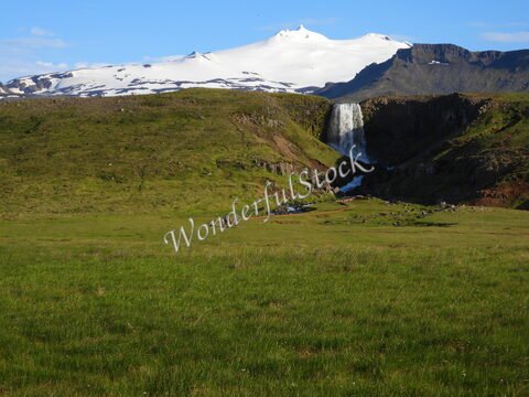 Chute d'eau à Svöðufoss, Islande, avec des montagnes enneigées en arrière-plan et des visiteurs sur un terrain verdoyant.