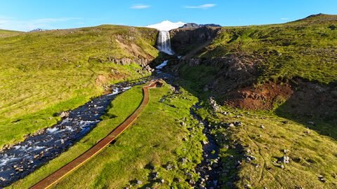 Svöðufoss en Islande, avec une chute d'eau entourée de collines verdoyantes et d'un chemin en bois le long d'un ruisseau.