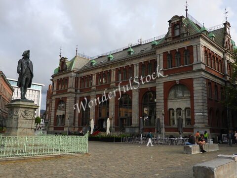 Statue de Holberg devant un bâtiment historique à Bergen, Norvège, avec des tables de café à l'extérieur et un ciel nuageux.