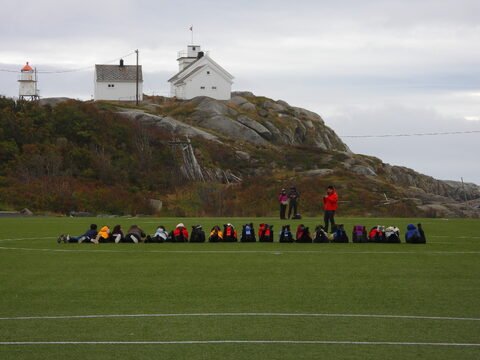 Des personnes allongées sur un terrain de sport à Henningsvær, Norvège, avec des maisons et un phare en arrière-plan.