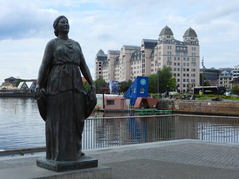 Statue d'une femme à Oslo, Norvège, avec des bâtiments modernes et un port en arrière-plan.