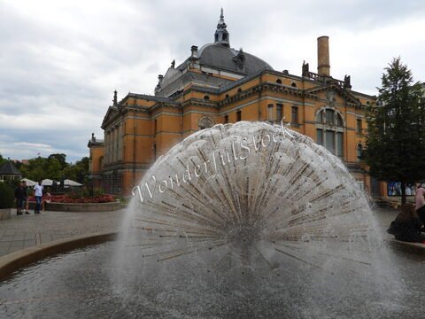 Fontaine en éventail devant un bâtiment historique à Oslo, Norvège, avec des fleurs et des arbres en arrière-plan.