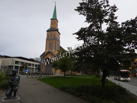 Église avec un clocher vert à Tromsø, Norvège, entourée de verdure, avec une sculpture et un bus en arrière-plan.