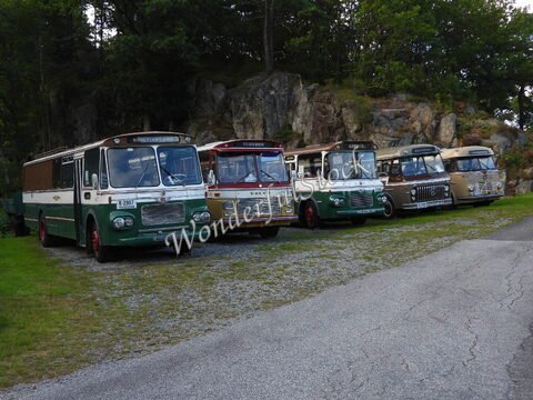 Un groupe de bus anciens est stationné sur une route à Kristiansand, Norvège, avec des rochers et des arbres en arrière-plan.