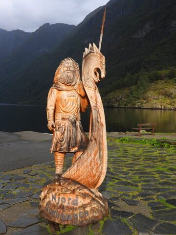 Statue en bois représentant Njord, à Gudvangen, Norvège, avec un paysage de montagnes et un fjord en arrière-plan.