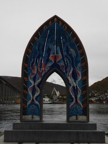 Monument coloré en mosaïque à Tromsø, Norvège, avec des montagnes en arrière-plan et un pont visible.