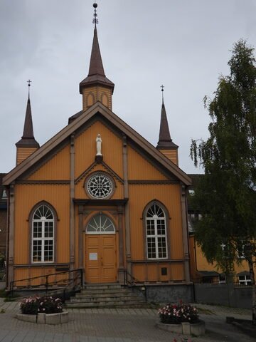 Église en bois avec des flèches, située à Tromsø, Norvège, entourée de fleurs et d'un ciel nuageux.