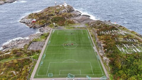 Terrain de football entouré par la mer à Henningsvær, Norvège, avec des maisons colorées et des rochers en arrière-plan.