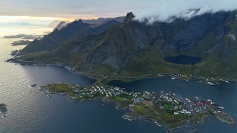 Vue aérienne d'un village côtier en Norvège, entouré de montagnes et d'une mer calme, avec des maisons colorées et des routes sinueuses.