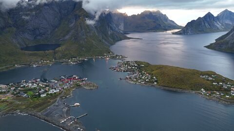 Vue aérienne d'un village côtier en Norvège, entouré de montagnes et d'un fjord, avec des maisons colorées et des bateaux dans l'eau.