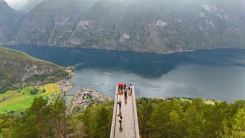Vue aérienne du point de vue Stegastein en Norvège, avec un lac et des montagnes en arrière-plan. Des visiteurs se tiennent sur la plateforme en bois.