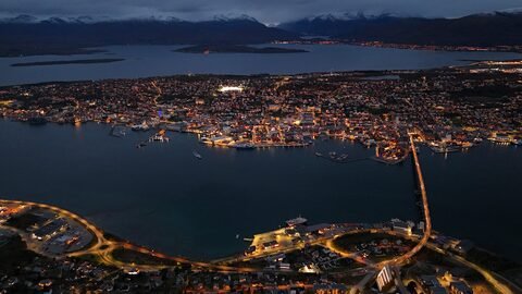 Tromsø, Norvège, vue aérienne d'une ville illuminée la nuit avec des montagnes en arrière-plan et un port visible.
