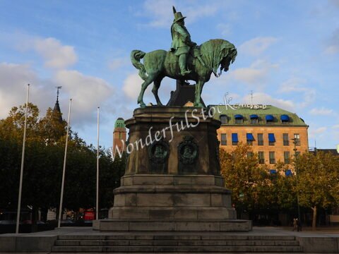Statue équestre en bronze à Malmö, Suède, avec des bâtiments modernes en arrière-plan et des arbres sur les côtés.