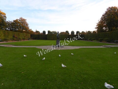 Un jardin avec une statue au centre, entouré de haies, à Malmö, Suède, avec des oiseaux sur la pelouse.