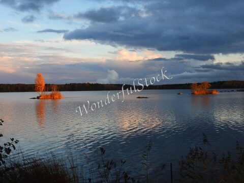 Un lac avec des îlots et des arbres, en Suède. Le ciel présente des nuages et des reflets sur l'eau au coucher du soleil.