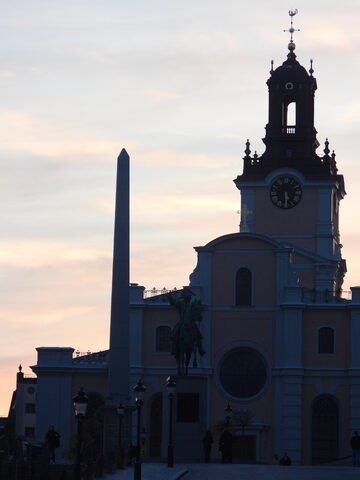 Édifice avec une horloge et une statue équestre à Stockholm, Suède, sous un ciel crépusculaire avec un obélisque en arrière-plan.