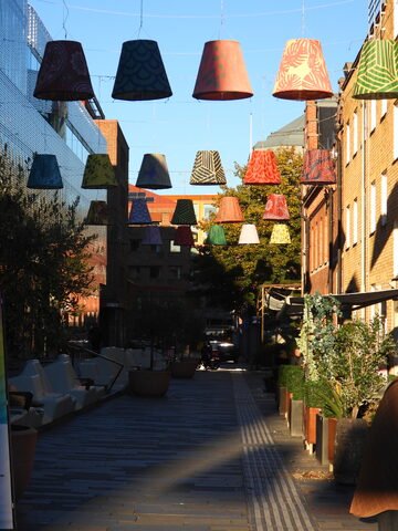 Des lampes colorées suspendues au-dessus d'une allée à Uppsala, Suède, entourées de végétation et d'architecture moderne.