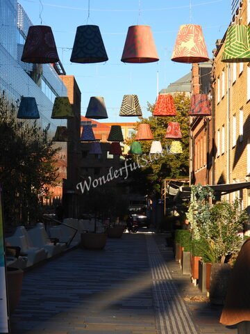 Des lampes colorées suspendues au-dessus d'une allée à Uppsala, Suède, entourées de végétation et d'architecture moderne.