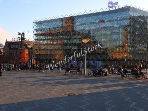 Bâtiment moderne en verre et pierre avec des enseignes, situé à Copenhague, Danemark, entouré de personnes et de bancs dans une place.
