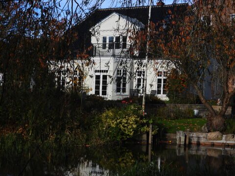 Maison blanche entourée de végétation à Ribe, Danemark, avec un reflet dans l'eau calme et des éléments de jardin visibles.
