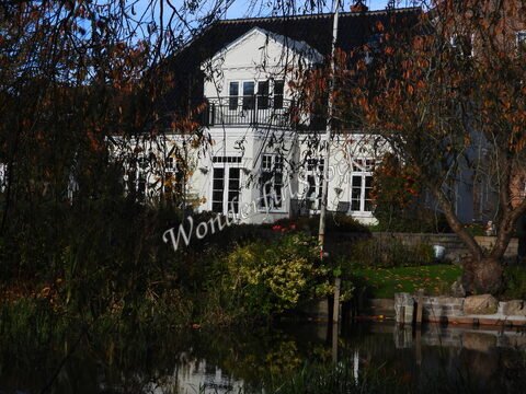 Maison blanche entourée de végétation à Ribe, Danemark, avec un reflet dans l'eau calme et des éléments de jardin visibles.