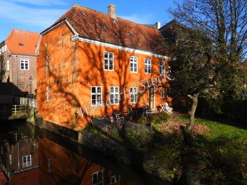Maison en briques orange avec fenêtres blanches et un jardin, située à Ribe, Danemark, reflétée dans un canal.
