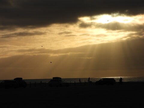 Plage à Romo, Danemark, avec des silhouettes de personnes et des voitures, sous un ciel nuageux laissant passer des rayons de lumière.