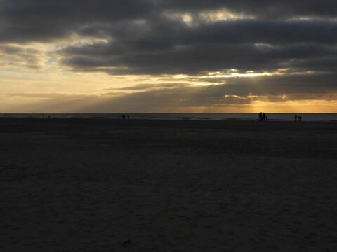 Plage de Romo, Danemark, avec des silhouettes de personnes se tenant près de l'eau sous un ciel nuageux et des rayons de soleil.