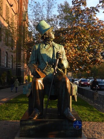 Statue d'H. C. Andersen à Copenhague, Danemark, entourée d'arbres et de bâtiments en brique, avec des passants et des véhicules en arrière-plan.