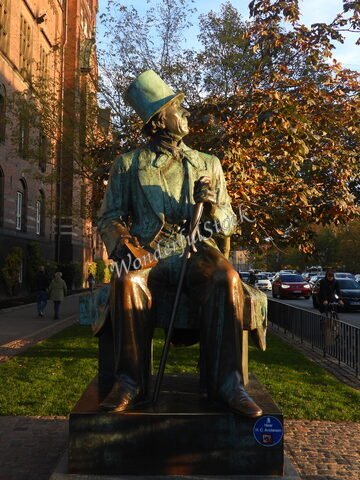 Statue d'H. C. Andersen à Copenhague, Danemark, entourée d'arbres et de bâtiments en brique, avec des passants et des véhicules en arrière-plan.