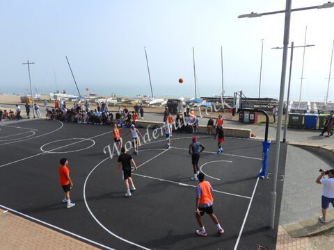 Match de basketball sur un terrain à Brighton, Angleterre, avec des joueurs et des spectateurs, en arrière-plan des bateaux.