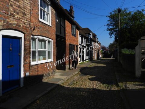 Des maisons en briques sur une rue pavée à Canterbury, en Angleterre, avec un ciel bleu et des câbles électriques visibles.