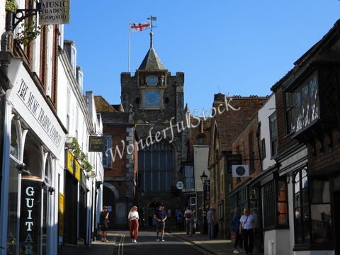 Rue animée à Canterbury, Angleterre, avec des bâtiments historiques et un drapeau anglais flottant au sommet d'une tour.