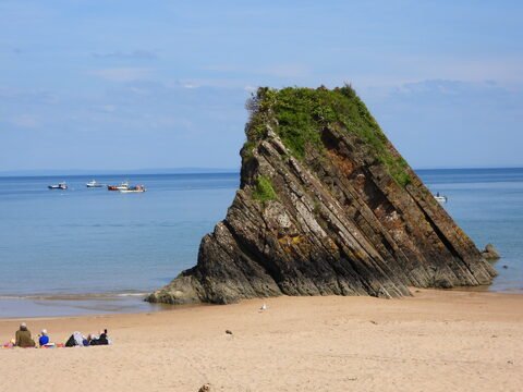 Rochers inclinés sur une plage de Tenby, au pays de Galles, avec quelques bateaux à l'horizon et des personnes assises sur le sable.