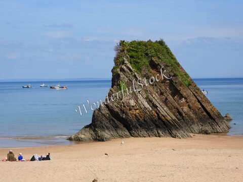 Rochers inclinés sur une plage de Tenby, au pays de Galles, avec quelques bateaux à l'horizon et des personnes assises sur le sable.